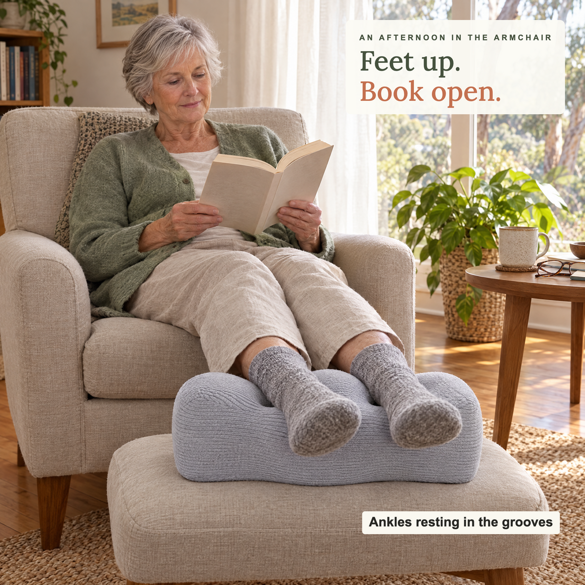 Woman reading in an armchair with her ankles resting in the grooves of the Comfort Leg Pillow on a footstool
