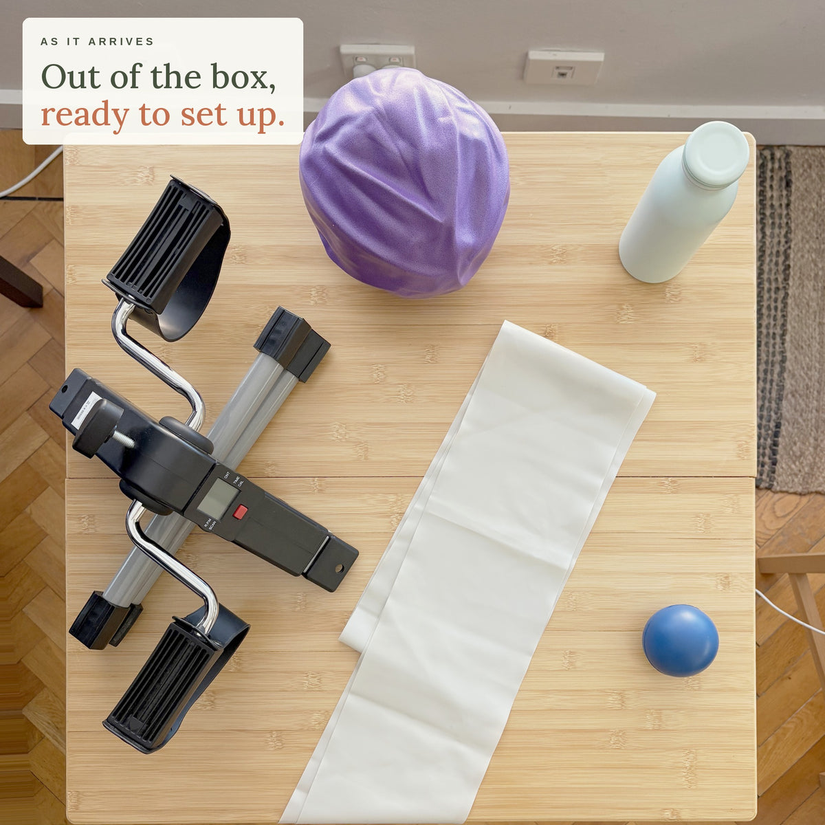 As it arrives. Out of the box, ready to set up: the Home Exercise Essentials Kit on a kitchen table seen from above, with the mini pedals, long resistance band, mini exercise ball, soft grip ball and bottle