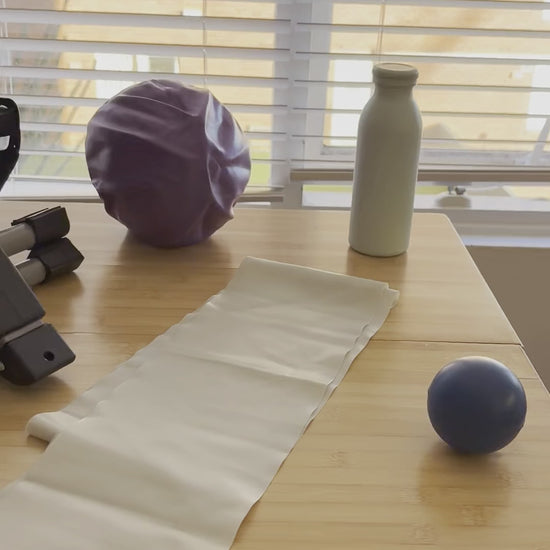 The Home Exercise Essentials Kit on a kitchen table at home: mini pedals, long resistance band, mini exercise ball, soft grip ball and bottle
