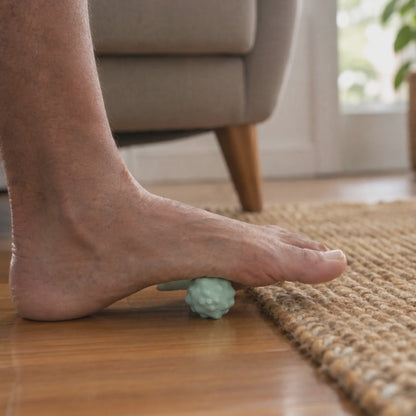 Side view: a bare foot rolling slowly heel to toe over the Peanut Massage Ball, which lies across the sole, in front of an armchair