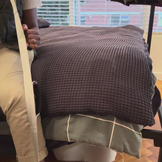 Sitting on the edge of the bed, the foot loop around the heel, using the Triple Grip Leg Lifter to guide the leg up onto the bed